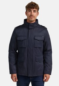 Marineblauwe jas met een hoge kraag, vier voorzakken, schouderepauletten en een gestructureerde afwerking. Voorzien van een knoopsluiting.