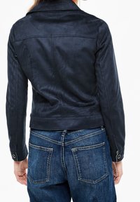 Giacca in suede blu navy con colletto, cuciture visibili e dettagli con bottoni a pressione, abbinata a jeans in denim blu con tasche posteriori.