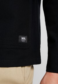 Svart tygjacka med ett synligt Vans-logomärke, med en robust design och strukturerad yta, i kombination med beige byxor.