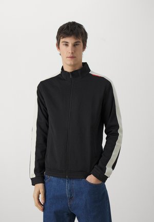 Giacca nera con zip e colletto alto, con strisce bianche laterali e logo rosso sulla spalla; tessuto morbido e design aderente.