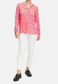 Femme portant une blouse longue à manches roses et orange à motifs, un pantalon blanc slim et des mocassins noirs brillants, se tenant sur un fond blanc.
