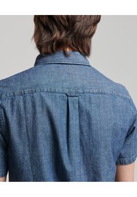 Koszula dżinsowa z krótkim rękawem w jasnoniebieskim kolorze, z tylną wkładką, subtelną fakturą i prostym kołnierzem. Tkanina wydaje się miękka i lekko wyprana.
