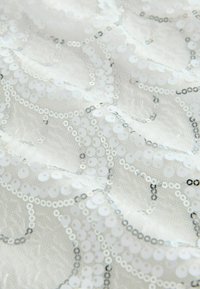 Tissu blanc transparent avec des motifs tourbillonnants de sequins translucides et argentés créant une surface texturée et scintillante.