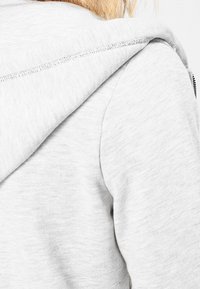 Sudadera de color gris claro con una tela suave y texturizada. Presenta una capucha distintiva y un detalle con cremallera en un lado. Costuras visibles.