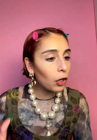 Jeune femme aux cheveux courts, barrettes colorées, grand collier de perles, boucles d'oreilles dorées et haut transparent à motif floral parle devant un fond rose.