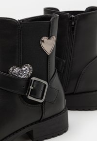 Černé kožené kotníkové boty s elastickým panelem, stříbrnou sponou a dvěma ozdobami ve tvaru srdce: jedna metalická a jedna třpytivá.