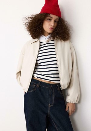 Junge Frau mit lockigem Haar, die eine rote Beanie, eine beigefarbene Jacke mit Reißverschluss, ein gestreiftes Shirt und dunkelblaue hoch taillierte Jeans trägt und vor einer weißen Wand steht.