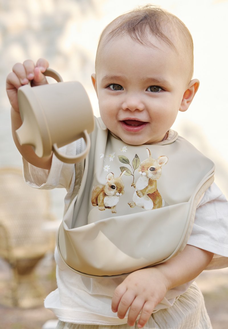 Silikonhaklapp i beige med kaninillustrationer, tillsammans med en beige mugg. Haklappen har en rundad design och mjuk textur, lämplig för barn.