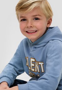 Jasnoniebieska bluza z kapturem z kieszenią typu kangur, z dużym białym i złotym napisem. Miękka tkanina o gładkiej teksturze i ściągaczami przy rękawach.