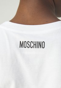 Biała bawełniana koszulka z czarnym logo "MOSCHINO" nadrukowanym na tylnej części przy kołnierzyku. Gładka faktura z klasycznym, okrągłym dekoltem.