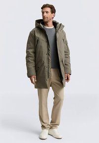 Oliivinvihreä parkatakki hupulla, edessä napitettava ja vetoketjullinen suljenta, sivutaskut, pidetty harmaan neuleen ja beigejen housujen päällä.