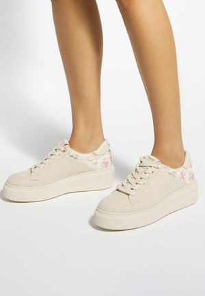 Feet wearing cream-colored platform sneakers with floral embroidery near the heel, standing on a white surface.