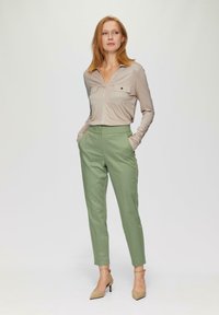 Beige Langarmshirt mit zwei Brusttaschen, kombiniert mit maßgeschneiderten hellgrünen Hosen. Model trägt nude-farbene High Heels, die Hände in den Taschen.