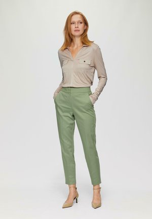 Beige blouse met lange mouwen en twee borstzakken, gecombineerd met op maat gemaakte lichtgroene broek. Model draagt naakte hakken, handen in de zakken.