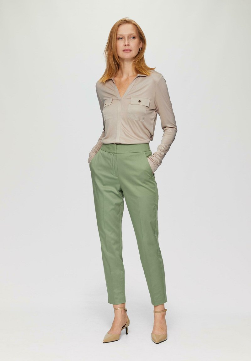 Beige Langarmshirt mit zwei Brusttaschen, kombiniert mit maßgeschneiderten hellgrünen Hosen. Model trägt nude-farbene High Heels, die Hände in den Taschen.