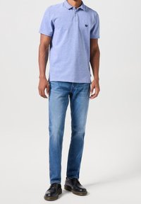 Hombre vistiendo una camiseta polo azul claro, jeans desgastados de color azul y zapatos de cuero negros, de pie contra un fondo liso.