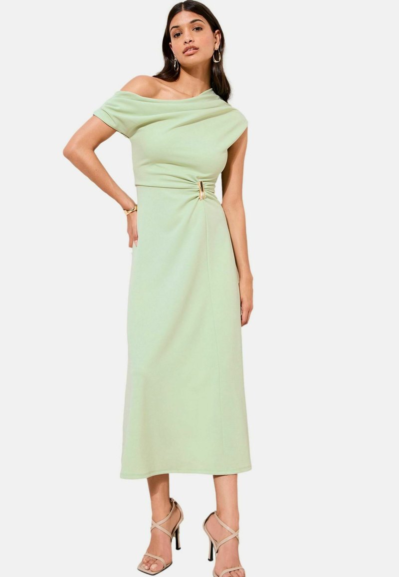 Žena nosi svetlo zelené midi šaty s odhalenými ramenami a zlatou prackou v páse, doplnené béžovými sandálmi na vysokom opätku.