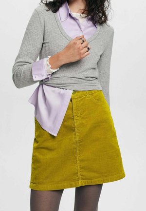 Femme portant une chemise lavande sous un pull gris côtelé avec des poignets volantés, associée à une jupe en velours côtelé jaune et des collants noirs.