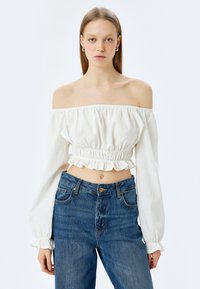 Hvid off-the-shoulder crop top med puffærmer og flæsekant, parret med højtaljede denim jeans med knaplukning og lommer.