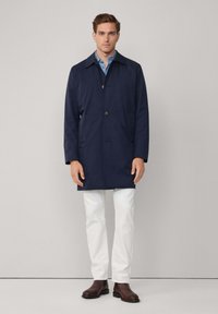 Cappotto blu navy, a lungo fino al ginocchio, realizzato in tessuto liscio, con chiusura a bottoni, ampio colletto e due tasche laterali, abbinato a pantaloni bianchi.