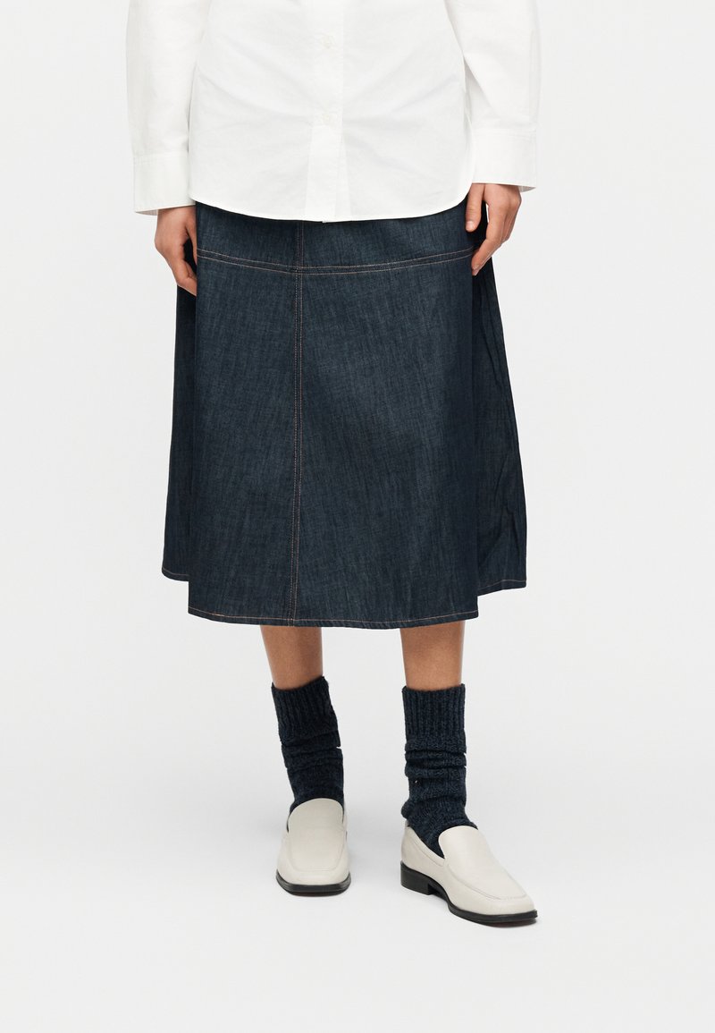 Άτομο που φοράει σκούρα τζιν φούστα μέχρι τα γόνατα, λευκό πουκάμισο με κουμπιά, μπλε κάλτσες πλεκτές και λευκά μοκασίνια, στέκεται μπροστά σε απλό φόντο.