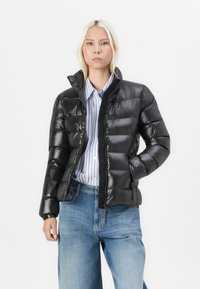 Kvinne med blondt hår iført en svart pufferjakke, stripete skjorte med knapper og løse blå jeans mot en ensfarget hvit bakgrunn.