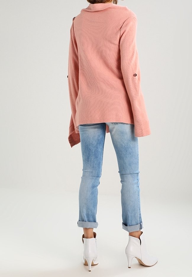 Femme portant un pull rose à manches longues, un jean bleu clair retroussé et des bottines blanches à talons, debout devant un fond uni.