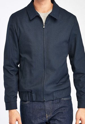 Homme portant une veste zippée bleu marine à fines rayures sur une chemise gris clair et un jean bleu foncé, debout les mains détendues le long du corps.