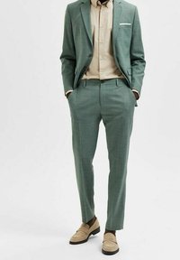 Man i skräddarsydd sagegrön kostym med beige skjorta och matchande beiga loafers, med händerna i fickorna, står framför en enkel vit bakgrund.