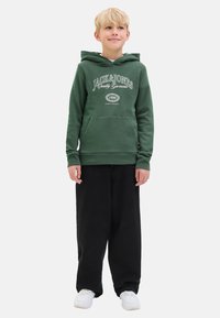 Hoodie vert avec une poche frontale et un logo imprimé blanc, associé à un pantalon noir ample et des baskets blanches. Look décontracté, tissu doux.