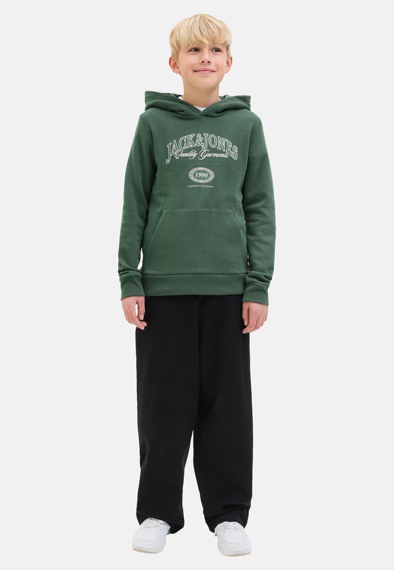 Hoodie vert avec une poche frontale et un logo imprimé blanc, associé à un pantalon noir ample et des baskets blanches. Look décontracté, tissu doux.