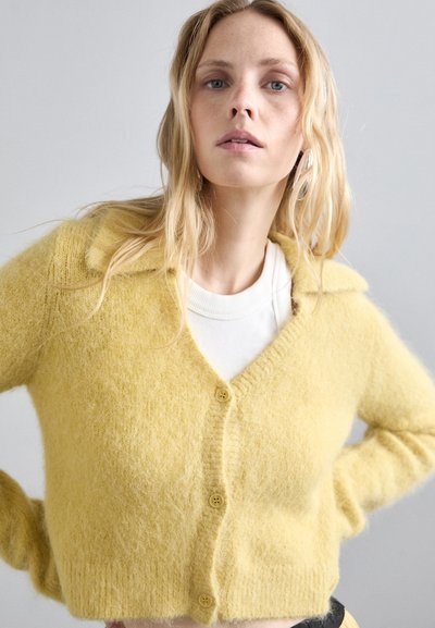 Κίτρινο, αφράτο ζακέτα με γιακά, ντεκολτέ και κούμπωμα με κουμπιά. Κοντό μήκος, μαλακή υφή, φοριέται πάνω από μια λευκή μπλούζα.