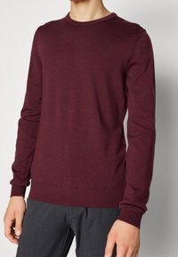 Homme portant un pull ajusté à manches longues bordeaux associé à un pantalon gris foncé, debout devant un fond clair uni.
