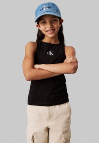 Canotta nera senza maniche con logo bianco "cK", abbinata a un cappellino in denim azzurro chiaro. Lo sfondo è di un grigio neutro.