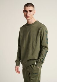 Olijfgroene sweatshirt met een ronde hals, ribbelmanchetten en een afgeronde zoom. Voorzien van contrasterende blauwe chevronstrepen op de mouwen.