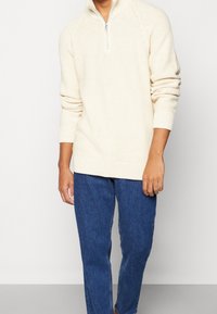Krémový pletený pulovr s límcem na půl zip a žebrovanými manžetami a lemem. Kombinován s modrými džínami pro ležérní vzhled.