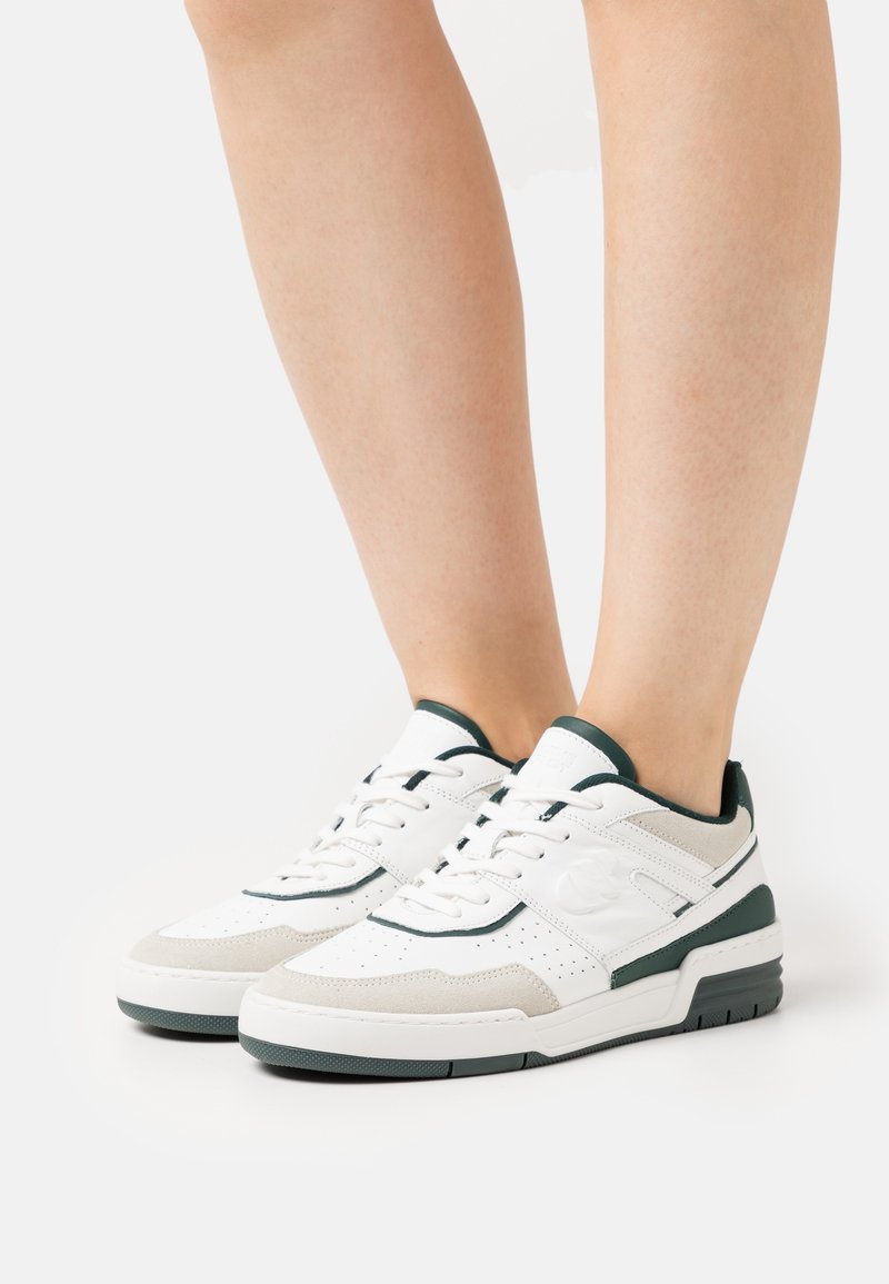 Personne portant des baskets basses blanches et grises avec un bord noir sur fond blanc, montrant les mollets et les pieds de côté.