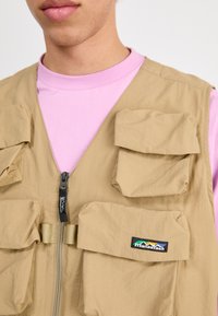 Beige Utility-Weste mit mehreren Fronttaschen, Reißverschluss und einem Logo-Patch. Über einem hellpinkfarbenen T-Shirt getragen. Glattes, leichtes Material.