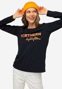 Granatowa koszulka z długim rękawem z pomarańczowym napisem "NORTHERN highlights". Model nosi jasnopomarańczową wełnianą czapkę, w zestawieniu z białymi spodniami.