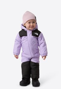Fialová zateplená zimní kombinéza s černými doplňky, předním zipem a elastickými manžetami. Kombinována s lehce růžovou pletenou čepicí a černými sněhovými botami.