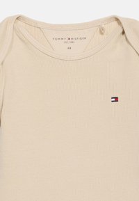 Μπεζ κορδωτό κορμάκι μωρού με λογότυπο Tommy Hilfiger στο στήθος και εκτυπωμένη ετικέτα εμπορικού σήματος στο εσωτερικό του λαιμού.