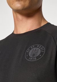 Svart T-skjorte med korte ermer og rund hals, med en grå FC St. Pauli-logo på venstre brystområde. Myk tekstilstruktur.