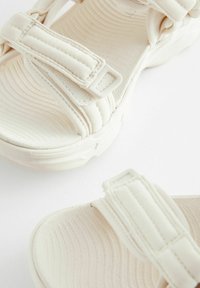 Sandales en tissu blanc avec des semelles texturées et des brides réglables. Elles présentent un dessus doux et matelassé ainsi que des boucles robustes pour le maintien.