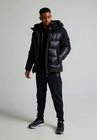 Chaqueta negra acolchada con capucha, con un acabado brillante y diseño acolchado. Combinada con pantalones jogger negros y zapatillas deportivas negras.