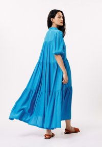 Blauwe katoenen maxi-jurk met gelaagd ontwerp en korte pofmouwen. Heeft een kraag en een vloeiende rok, gedragen met tan platte sandalen.