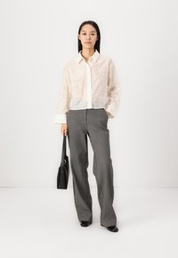 White lace blouse with a button-down design, paired with high-waisted striped gray trousers. Model holds a black handbag.