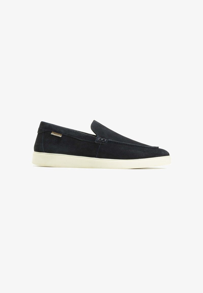 Chaussures slip-on en daim noir au design minimaliste, semelle en caoutchouc blanc et coutures d'accent. Présente une petite étiquette de marque sur le côté.