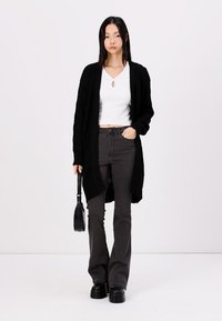 Cardigan nero a maglia sopra una maglietta crop bianca, abbinato a jeans svasati scuri e scarpe nere chunky. Modella con una piccola borsa a tracolla nera.