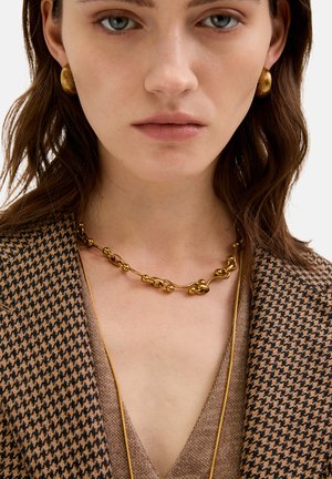 Złoty naszyjnik w postaci łańcuszka z powiązanymi owalnymi kształtami i kulistymi akcentami, w zestawieniu z marynarką w wzór hounds-tooth. Gładka tekstura.