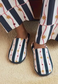 Pantofole pelose con motivi a stella blu, bianchi e dorati, caratterizzate da una texture morbida e un design arrotondato, indossate con pantaloni del pigiama a righe.